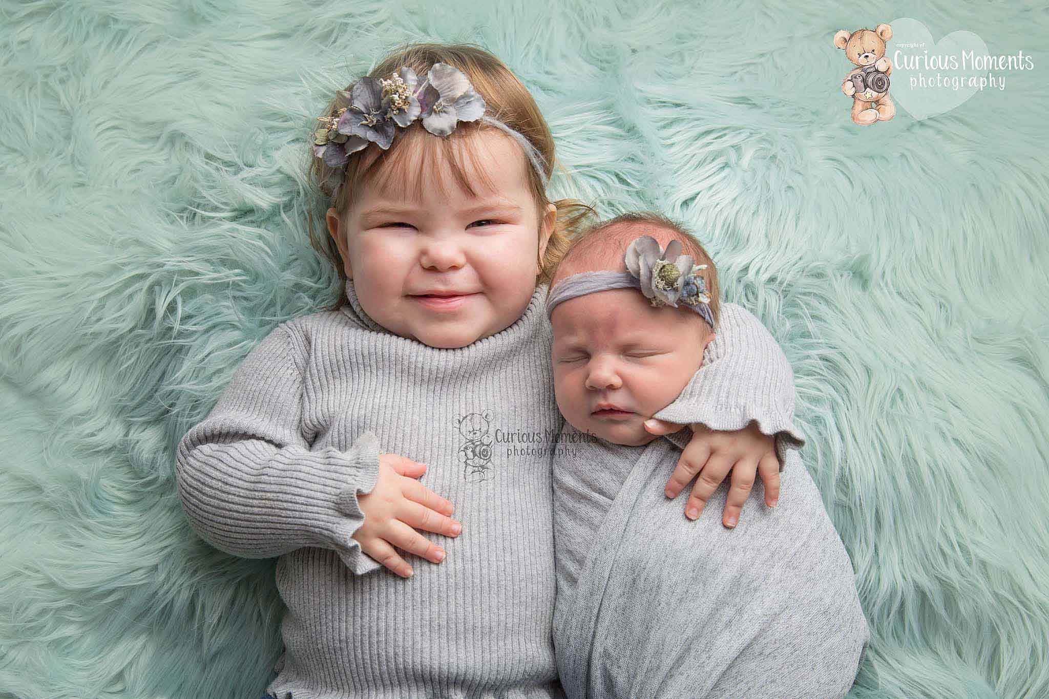 newborn baby girl wrapped in light grey being snuggled by older sister on a mint green background