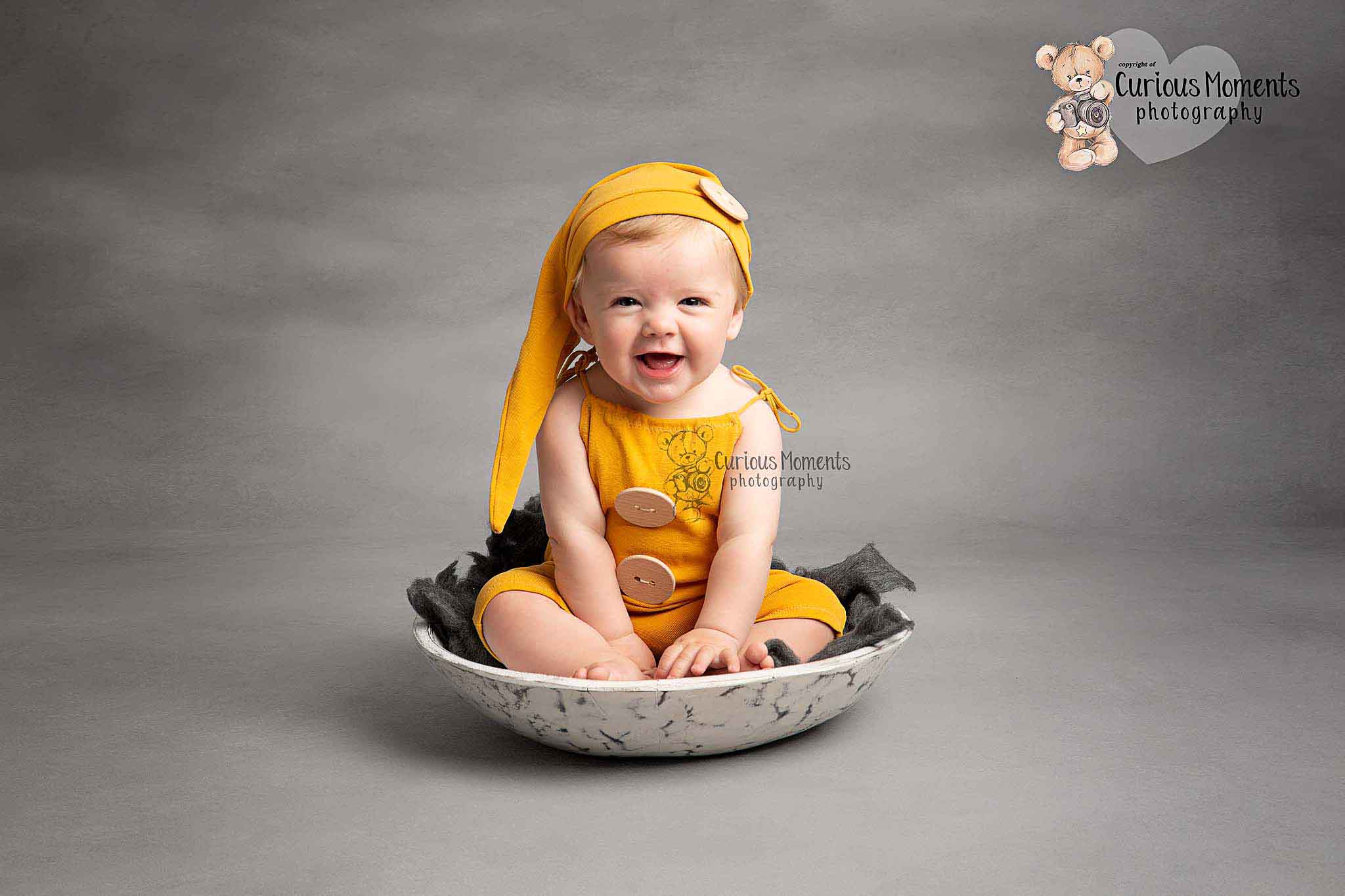 Baby bow wearning yellow romper and sleepy hat sat in a white mottled bowl on a grey background